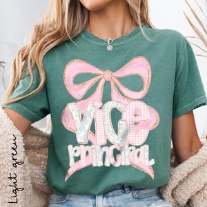 May include: Light green t-shirt with a pink bow and the words "VICE PRINCIPAL" in a mix of floral and gingham patterns. The shirt has short sleeves and a crew neck.