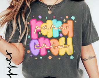 May include: Dark gray t-shirt with the words "Para Crew" in colorful bubble letters. The text is outlined in white and accented with small flower and star graphics. The shirt is a casual style.