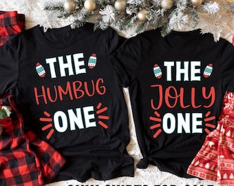 May include: Two black t-shirts with a Christmas theme. The left shirt says "The Humbug One" with a red and white bauble graphic. The right shirt says "The Jolly One" with a red and white bauble graphic. The shirts are on a white background with red and white plaid fabric and Christmas decorations.