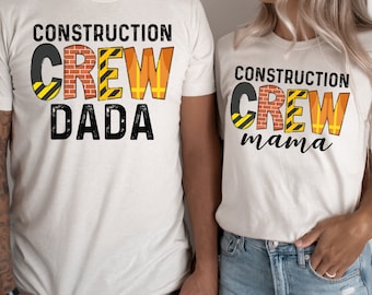 Peut inclure: Deux t-shirts blancs avec le texte "Construction Crew Dada" et "Construction Crew Mama" en noir et jaune. Le texte est stylisé avec des images de briques, de cônes de chantier et de ruban adhésif de signalisation.