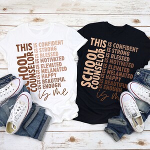 May include: Two t-shirts, one white and one black, with the words "School Counselor" and inspirational phrases printed in brown. White sneakers and folded blue jeans are placed near the shirts.