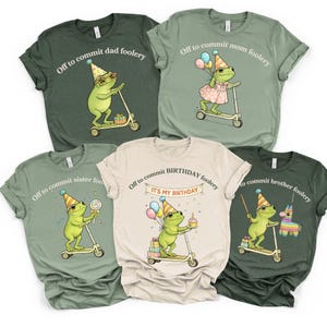 May include: A collection of five t-shirts in various colors, each featuring a cartoon frog riding a scooter. The shirts have phrases like "Off to commit dad foolery" and "Off to commit birthday foolery". The frog illustrations are whimsical and colorful.