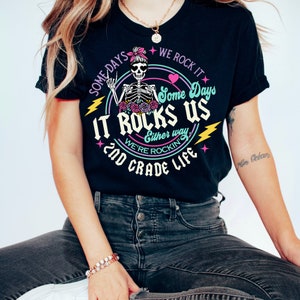 May include: Black t-shirt with a graphic of a skeleton wearing a pink bow and holding a peace sign. The graphic also includes the text "Some Days We Rock It Some Days It Rocks Us Either Way We're Rockin' 2nd Grade Life".