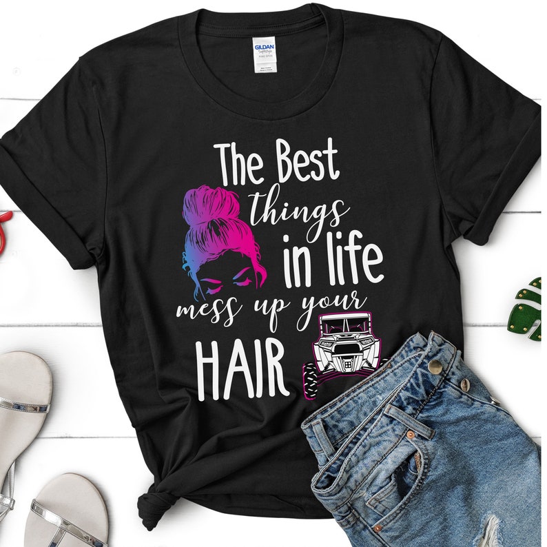 May include: Black t-shirt with a graphic design featuring a woman with a messy bun and a quote that reads "The best things in life mess up your hair". The graphic also includes a silhouette of an off-road vehicle.