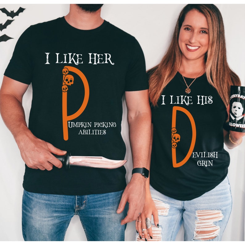 May include: Two black t-shirts with orange text and graphics. The first shirt says "I like her" with a stylized orange "D" with skulls and the text "Pumpkin Picking Abilities". The second shirt says "I like his" with a stylized orange "D" with skulls and the text "Evilish Grin".
