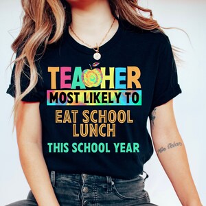 以下が含まれることがあります： 「Teacher most likely to eat school lunch this school year」というテキストが書かれたタイダイ柄の黒いTシャツ。テキストは虹色で、単語「Teacher」にはタイダイ柄のリンゴが描かれています。