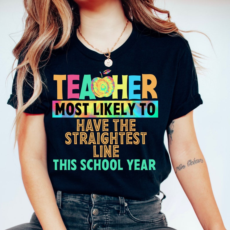以下が含まれることがあります： 「Teacher most likely to have the straightest line this school year」というテキストが書かれたタイダイ柄の黒いTシャツ。デザインには虹色のリンゴが含まれています。