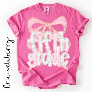 May include: A bright pink short-sleeve t-shirt featuring a large graphic of a light pink bow with gold glitter outlines. The text "fifth grade" is printed in a mix of light pink gingham and blue floral patterns with gold glitter outlines.