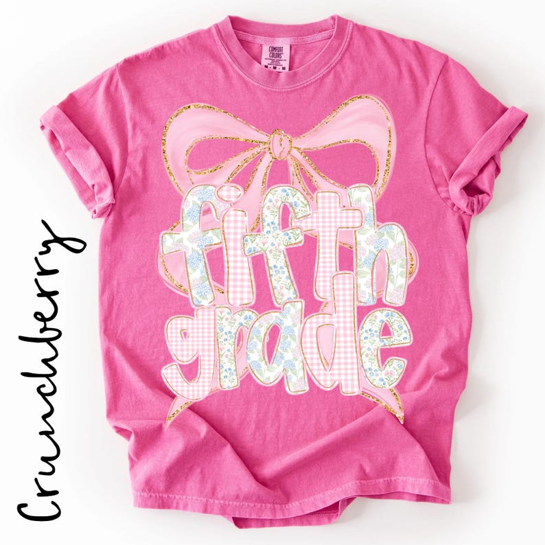 May include: A bright pink short-sleeve t-shirt featuring a large graphic of a light pink bow with gold glitter outlines. The text "fifth grade" is printed in a mix of light pink gingham and blue floral patterns with gold glitter outlines.