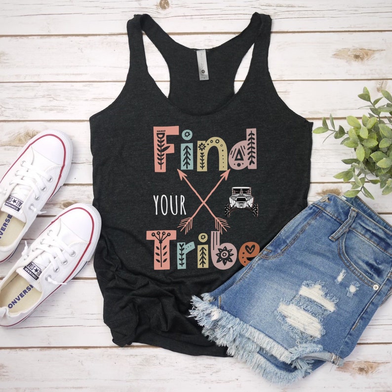 May include: Black tank top with the text "Find Your Tribe" in colourful letters with a graphic of crossed arrows and a jeep. The tank top is lying on a white wooden surface with a pair of white sneakers and a pair of blue denim shorts.