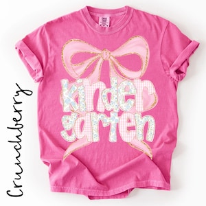 Peut inclure: Un t-shirt rose vif à manches courtes orné d'un grand nœud rose clair avec des contours dorés pailletés. Le mot kindergarten est imprimé avec des motifs floraux et vichy roses. Le texte Crunchberry apparaît verticalement sur la gauche.