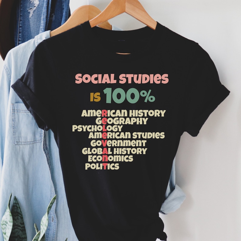 Op de afbeelding: Zwart T-shirt met de tekst "SOCIAL STUDIES IS 100%" en een lijst met vakken in een kleurrijk, retro lettertype. De vakken omvatten Amerikaanse geschiedenis, geografie, psychologie, Amerikaanse studies, overheid, wereldgeschiedenis, economie en politiek.