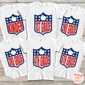 May include: Five white t-shirts with a blue and red shield design featuring a red apple and the text "Kinder", "1st Grade", "2nd Grade", "3rd Grade", and "4th Grade".