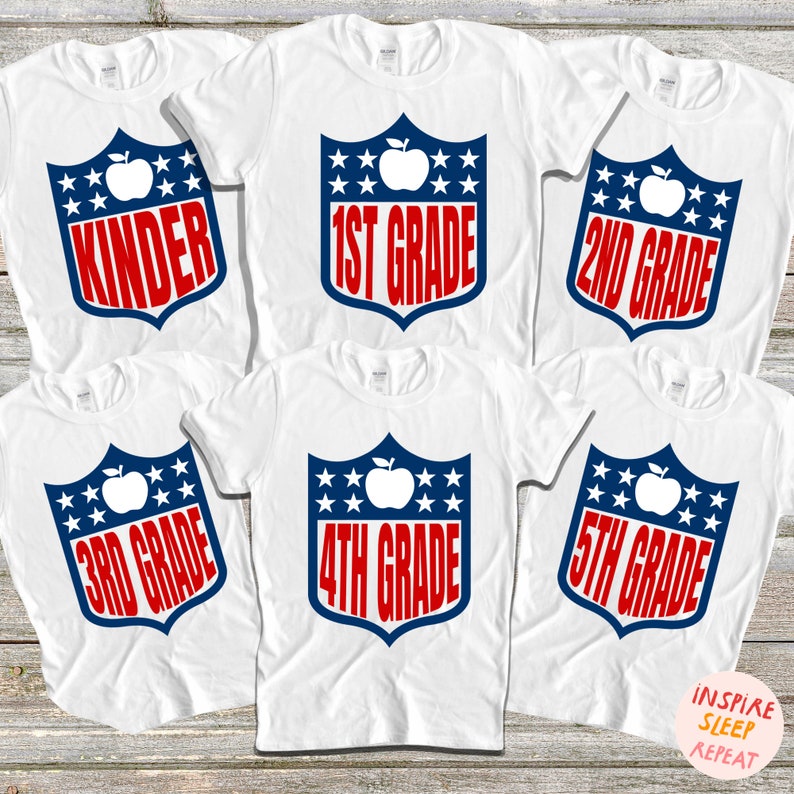 May include: Five white t-shirts with a blue and red shield design featuring a red apple and the text "Kinder", "1st Grade", "2nd Grade", "3rd Grade", and "4th Grade".