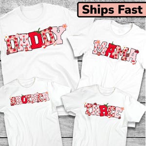 May include: Four white t-shirts with short sleeves. Each shirt has a different word in a strawberry-themed design: "Daddy", "Mama", "Brother", and "Three". The text is in red, pink, and white, with strawberry and floral accents.