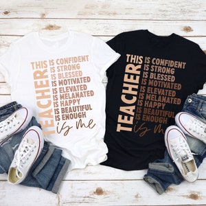 Puede incluir: Dos camisetas con el texto "This is confident, is strong, is blessed, is motivated, is elevated, is melanated, is happy, is beautiful, is enough, is me" impreso en ellas. El texto est&aacute; en una columna vertical y es de color marr&oacute;n y melocot&oacute;n. Las camisetas est&aacute;n sobre una superficie de madera con vaqueros azules y zapatillas blancas.