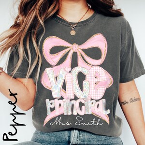 May include: Dark gray t-shirt with a pink bow and the words "VICE PRINCIPAL" in a mix of floral and gingham patterns. The name "Mrs. Smith" is written below. The shirt has a casual, vintage look.