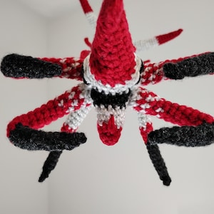 May include: A handmade crochet creature with a red, black, and white color scheme. The creature has a conical head, multiple arms, and black tips. The crochet work is detailed, showcasing a unique design.