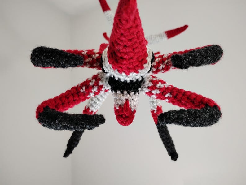 May include: A handmade crochet creature with a red, black, and white color scheme. The creature has a conical head, multiple arms, and black tips. The crochet work is detailed, showcasing a unique design.