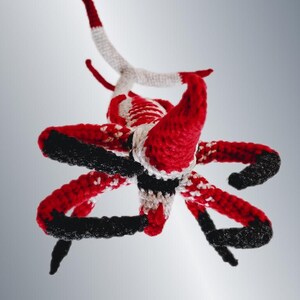 May include: A handmade crochet sculpture in red, black, and white. The sculpture has multiple curved arms and horns, with a central body. The color scheme is consistent throughout the piece, with the red and black dominating the design.