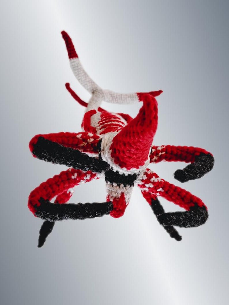 May include: A handmade crochet sculpture in red, black, and white. The sculpture has multiple curved arms and horns, with a central body. The color scheme is consistent throughout the piece, with the red and black dominating the design.