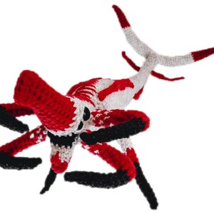May include: A handmade crochet creature in red, white, and black. The toy has multiple legs and a tail, with a detailed design. The creature's body is primarily red and white, with black accents on the legs and head.