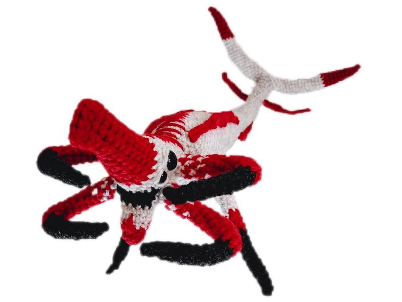 May include: A handmade crochet creature in red, white, and black. The toy has multiple legs and a tail, with a detailed design. The creature's body is primarily red and white, with black accents on the legs and head.