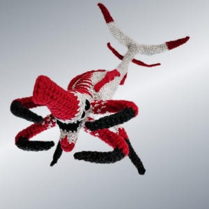 May include: A handmade crochet creature in red, white, and black. The toy has a large red head, black and red arms, and a white tail with red tips. The creature is displayed against a gray background.