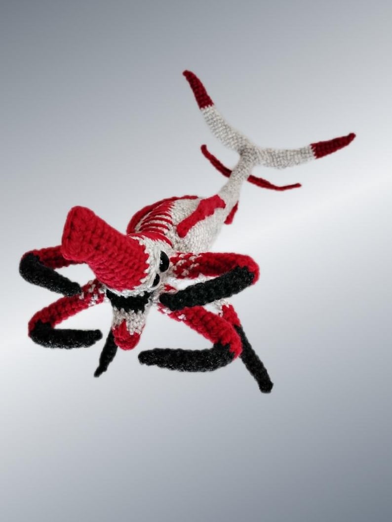 May include: A handmade crochet creature in red, white, and black. The toy has a large red head, black and red arms, and a white tail with red tips. The creature is displayed against a gray background.
