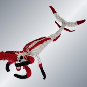 May include: A handmade crochet creature in red, white, and black. The whimsical design features a long body with red stripes, a red head, and black curved arms. The creature's tail and feet are tipped with red.