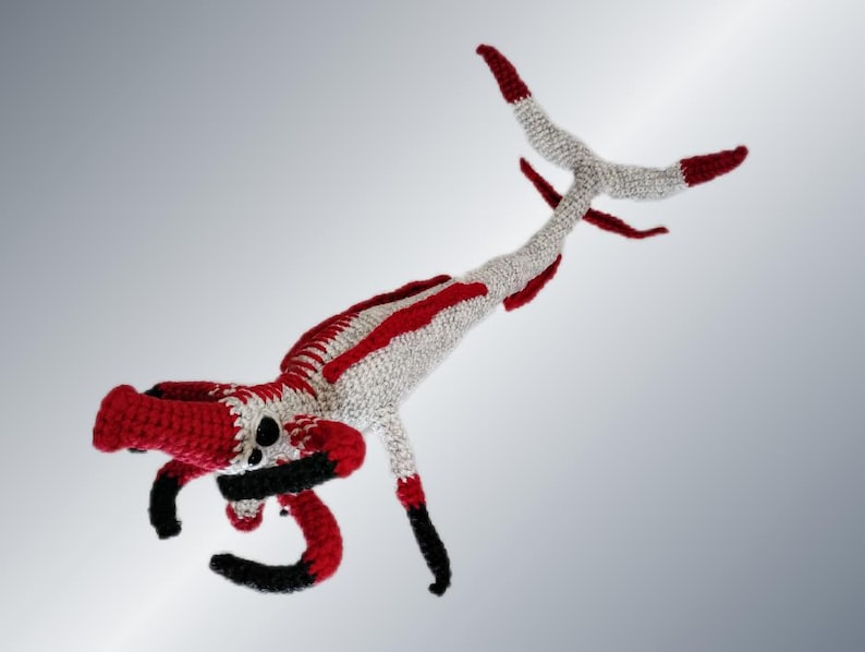 May include: A handmade crochet creature in red, white, and black. The whimsical design features a long body with red stripes, a red head, and black curved arms. The creature's tail and feet are tipped with red.