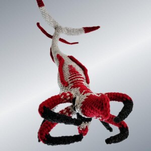 May include: A handmade crochet creature in red, black, and gray. The whimsical design features multiple arms and a long, segmented tail. The creature's body is primarily red and gray, with black accents on the arms and tail tips.