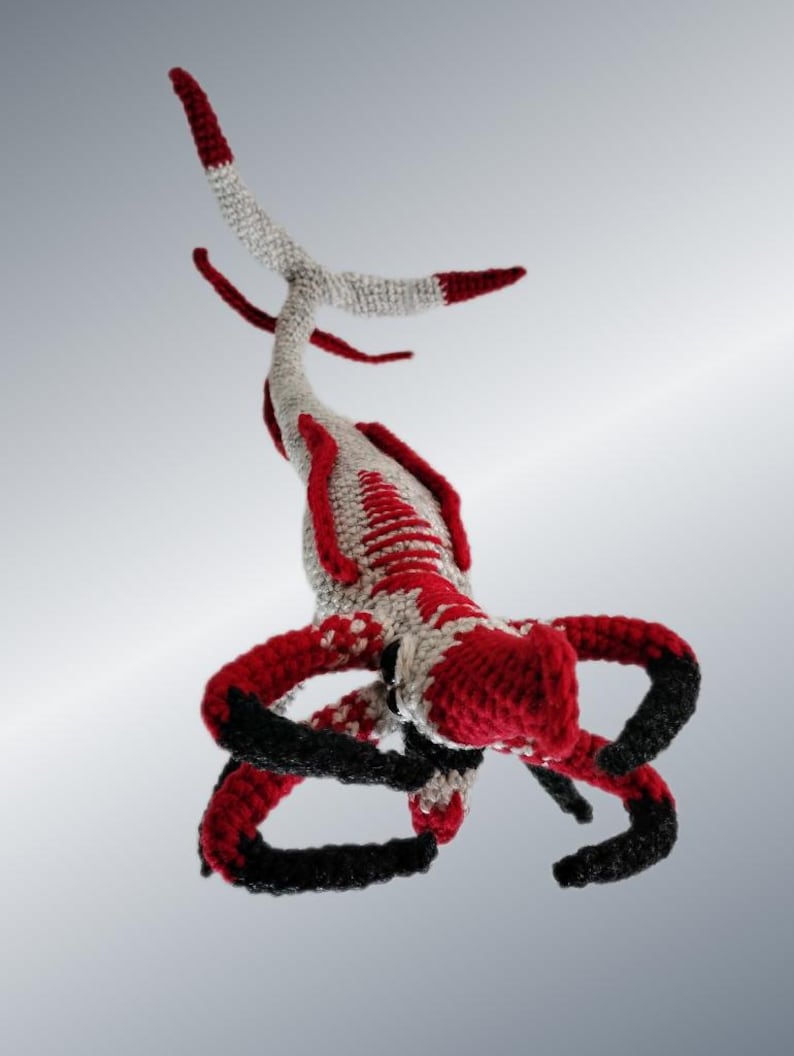 May include: A handmade crochet creature in red, black, and gray. The whimsical design features multiple arms and a long, segmented tail. The creature's body is primarily red and gray, with black accents on the arms and tail tips.