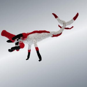 May include: A handmade crochet dragon in white, red, and black. The dragon has a long, curved body with a red head and tail tips. Its legs are black with red accents, and it appears to be a whimsical, fantasy-themed item.