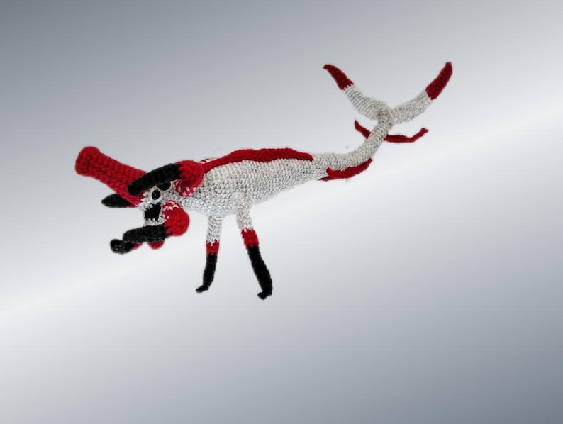 May include: A handmade crochet dragon in white, red, and black. The dragon has a long, curved body with a red head and tail tips. Its legs are black with red accents, and it appears to be a whimsical, fantasy-themed item.