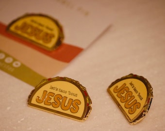 May include: Gold-colored enamel pins in the shape of a taco shell. The pins have the text "let's taco 'bout JESUS" in a brown font. One pin is on a card with a green and orange stripe.