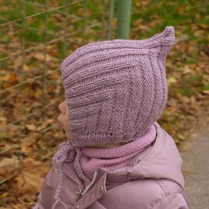 Könnte beinhalten: Eine gestrickte lavendelfarbene Mütze mit spitzer Spitze, getragen von einem Kind. Die Mütze hat ein strukturiertes, geripptes Design und wird mit einem passenden lavendelfarbenen Schal kombiniert. Das Kind trägt eine rosa Jacke.