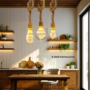 Peut inclure: Trois lampes suspendues avec des ampoules Edison sont suspendues à des cordes nouées dans une cuisine. Les ampoules sont allumées, dégageant une lueur chaleureuse. La cuisine comprend des meubles en bois et une table à manger. Le texte "© 2025 KitchenJoint" est visible.