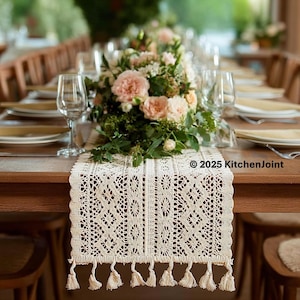 Puede incluir: Camino de mesa de encaje color crema con un patrón geométrico y flecos de borlas. Se coloca sobre una mesa de madera preparada para un evento formal, con arreglos florales y cristalería. El texto "© 2025 KitchenJoint" es visible.