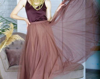 May include: A woman wearing a brown tulle skirt with a brown satin top and a gold layered necklace. She is standing in front of a white screen and a chair.