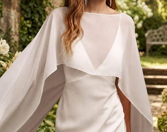 May include: A floor-length white gown featuring a sleeveless bodice with a V-neckline and a sheer, flowing cape overlay that drapes over the shoulders. The garment is crafted from a smooth, solid fabric and includes long, translucent cape panels.