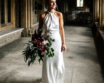 May include: A bride in a white halter-neck wedding dress holds a bouquet of dark red, pink, and green flowers in a stone hallway.
