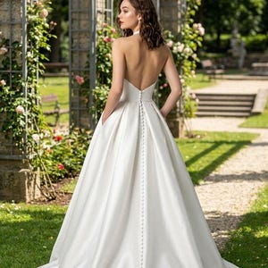 May include: A floor-length white satin wedding dress featuring a backless design, a halter neckline, and a long row of fabric-covered buttons running down the center back. The gown has a full, pleated skirt with integrated side pockets.