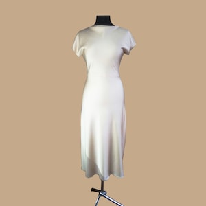 May include: A simple, off-white midi dress with short sleeves and a slightly A-line silhouette. The dress is made of a smooth, silky fabric.