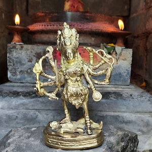 May include: A gold-colored metal statue of a deity with multiple arms, holding various objects. The statue is standing on a base with intricate details. The statue is in front of a brick wall with two lit candles.