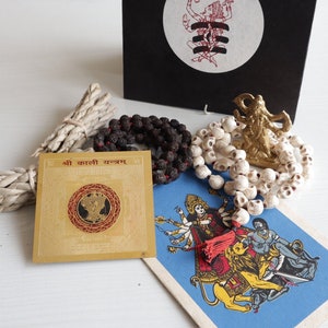 May include: A set of Hindu religious items including a gold yantra, a white skull mala, a black bead mala, a small gold statue of Kali, and a card with a colourful image of Kali. The card has the text "Shri Kali Yantra" in red and black.