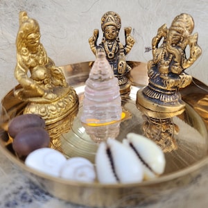 Peut inclure: Un plateau en laiton avec quatre petites statues dorées de divinités hindoues, une pyramide de cristal clair et des coquillages blancs.