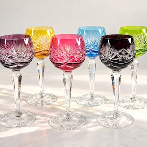 Op de afbeelding: Een set van zes kristallen wijnglazen in levendige kleuren, waaronder paars, geel, rood, blauw, donkerpaars en groen. Elk glas heeft een heldere, gefacetteerde steel en voet met een gedetailleerd stervormig slijppatroon in de gekleurde kelk.