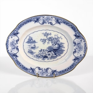 Op de afbeelding: Een ovale keramische serveerschaal met een blauw-wit transferpatroon. Het midden toont een landschap met een vaas met bloemen, een klein bootje en een pagode in de verte. De geschulpte rand heeft een gouden rand en florale krulmotieven.