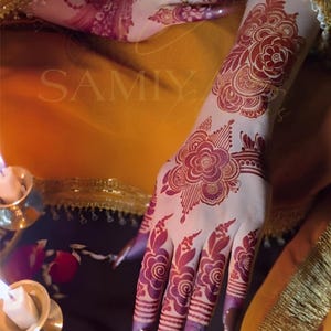 May include: Close-up of a hand adorned with intricate henna designs. The henna is a deep reddish-brown color, featuring floral patterns on the wrist and fingers. The nails are painted a dark brown. The background is a mix of orange and gold.